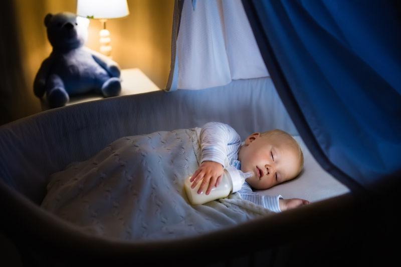 可爱的婴儿喝牛奶在蓝色的摇篮与雨篷在夜间