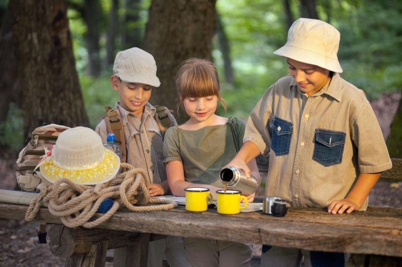 森林里站在木桌旁正在倒水喝的野外探险的孩子们