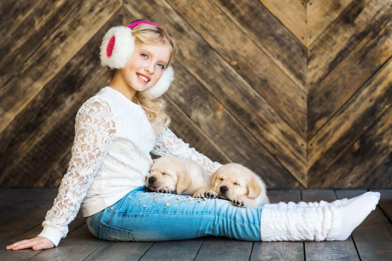 木背景下坐在地上抱着拉布拉多小狗的金发小女孩