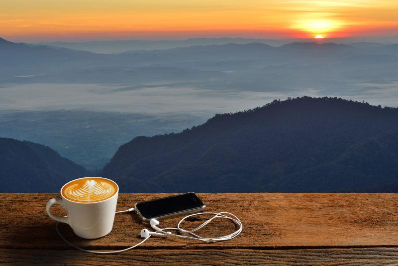 日出时分木桌上的一杯热咖啡和一只带着耳机的智能手机