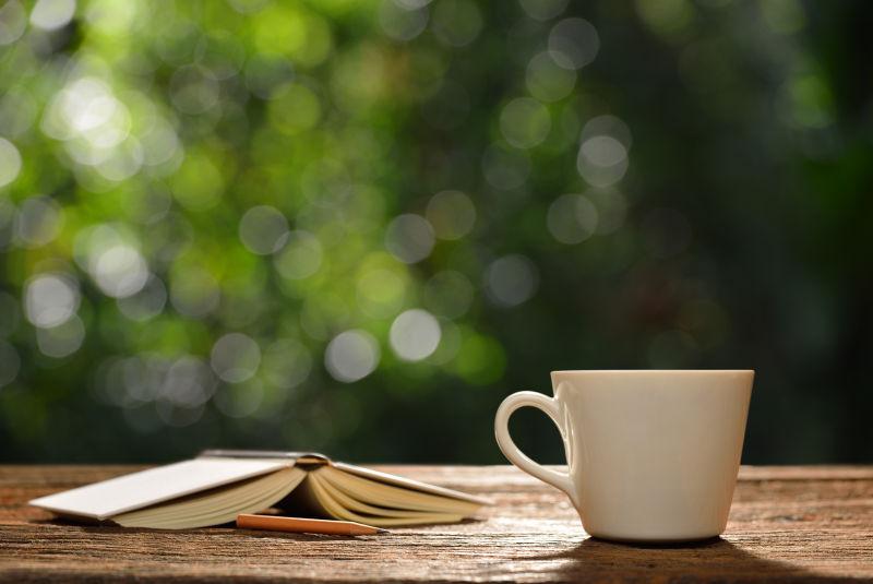 绿色户外阳光下的木桌面上的一本书和一杯热咖啡