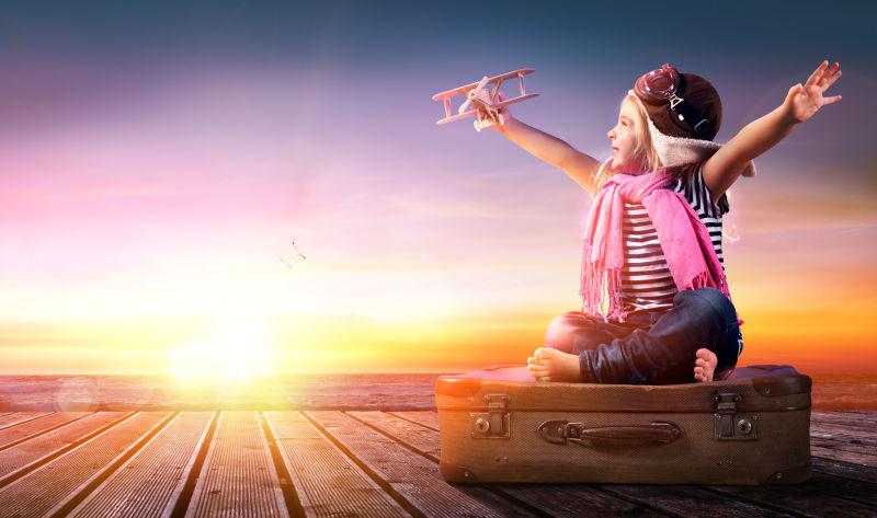 夕阳下坐在行李箱上玩玩具飞机的小女孩