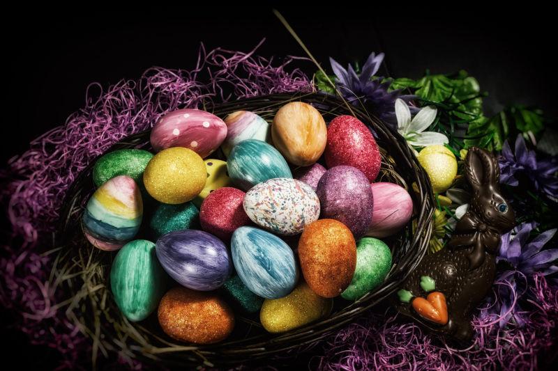 黑色背景下紫色草窝里的篮子里的复活节彩蛋和巧克力兔子