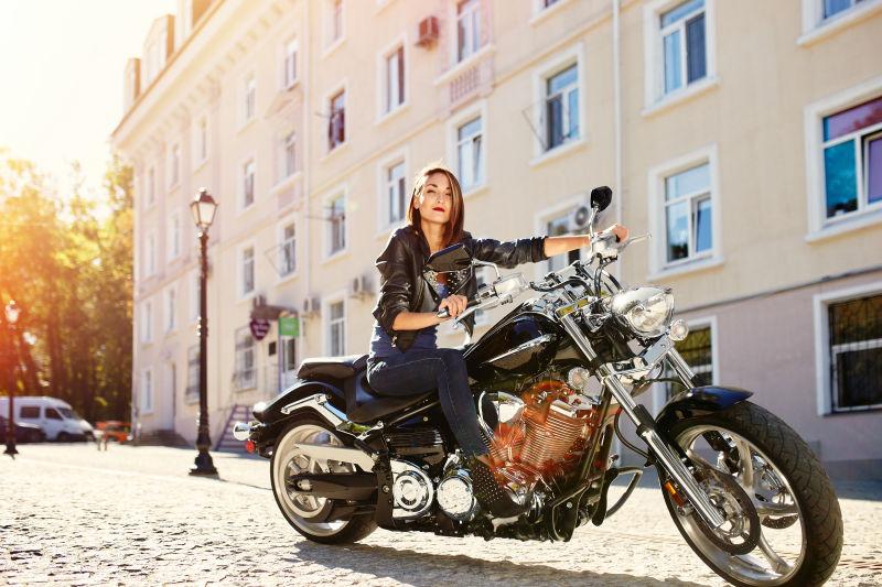 阳光下的建筑前穿着黑色皮夹克坐在摩托车上的美女