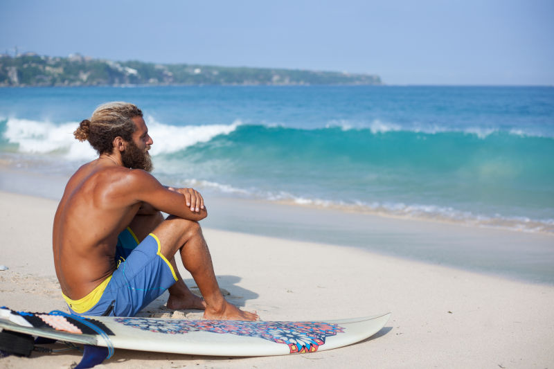 蹲坐在海滩上的男人身边放着冲浪板
