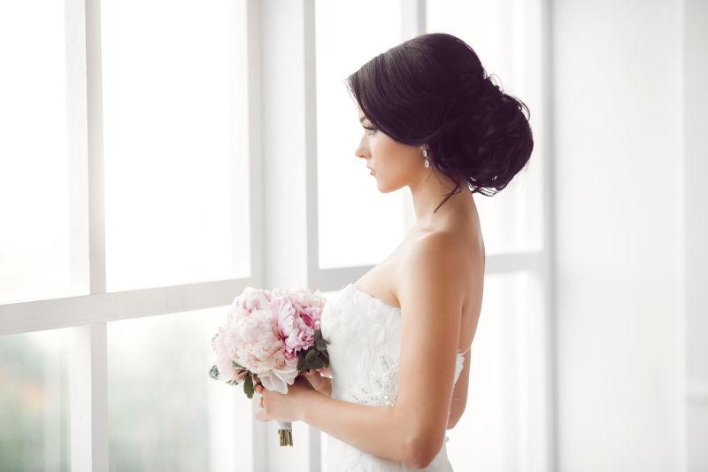 拿手捧花的美丽新娘站在窗前
