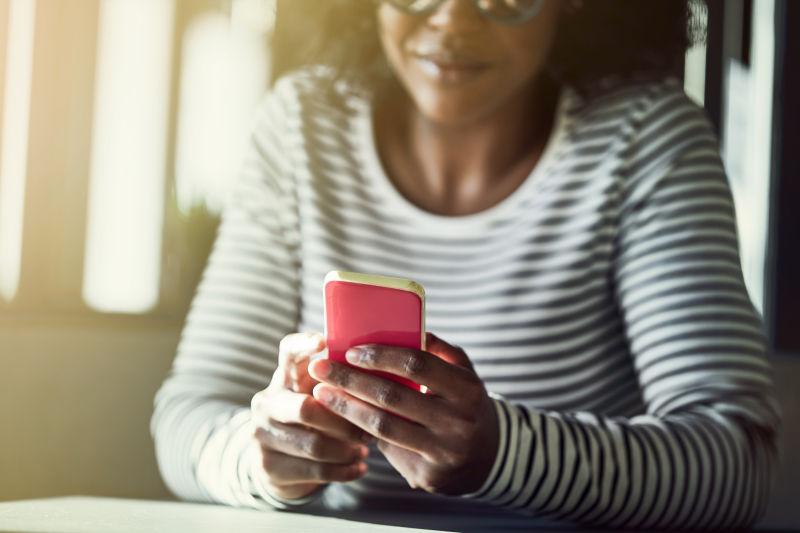 戴着眼镜的年轻非洲妇女微笑着坐在桌旁阅读手机上的短信