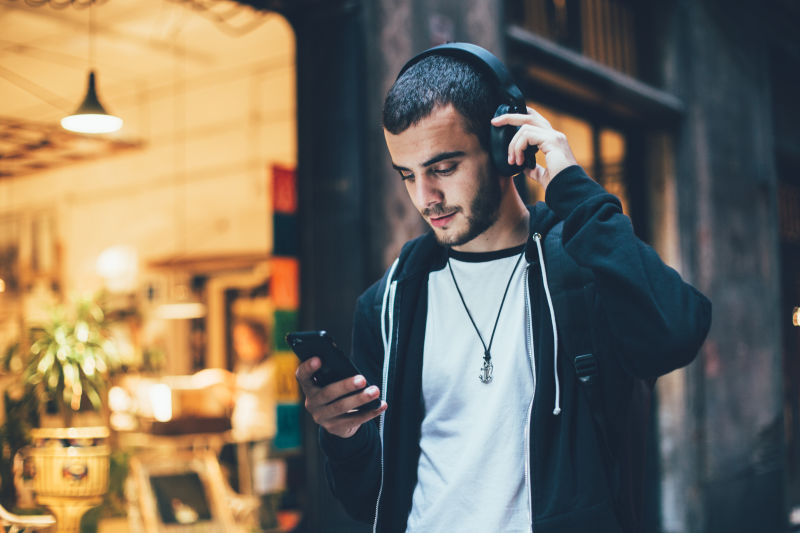 西班牙裔年轻有魅力的男人站在黑暗的街道前在智能手机上变换歌曲和音轨用无线耳机听音乐有轻微胡须的嬉皮士