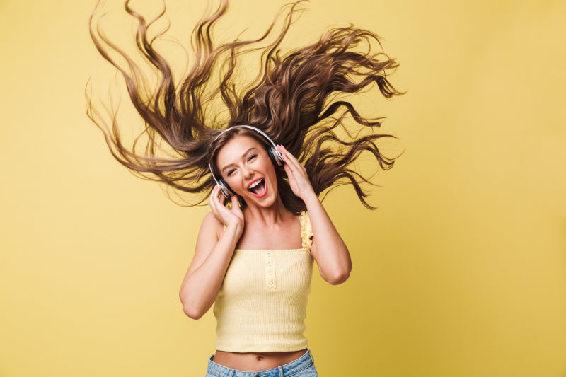 通过耳机收听音乐时20多岁的有趣女性在唱歌和享受晃动头发的乐趣背景为黄色