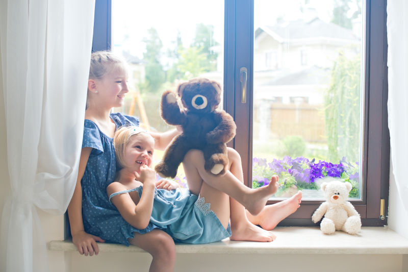 两个可爱的欧洲小女孩坐在家里靠窗的地方玩泰迪熊