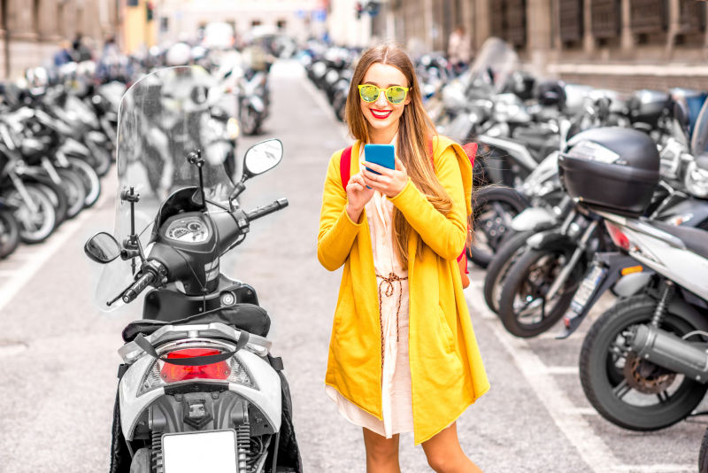 穿着黄色毛衣的年轻女士在意大利的街道上使用满是滑板车的手机