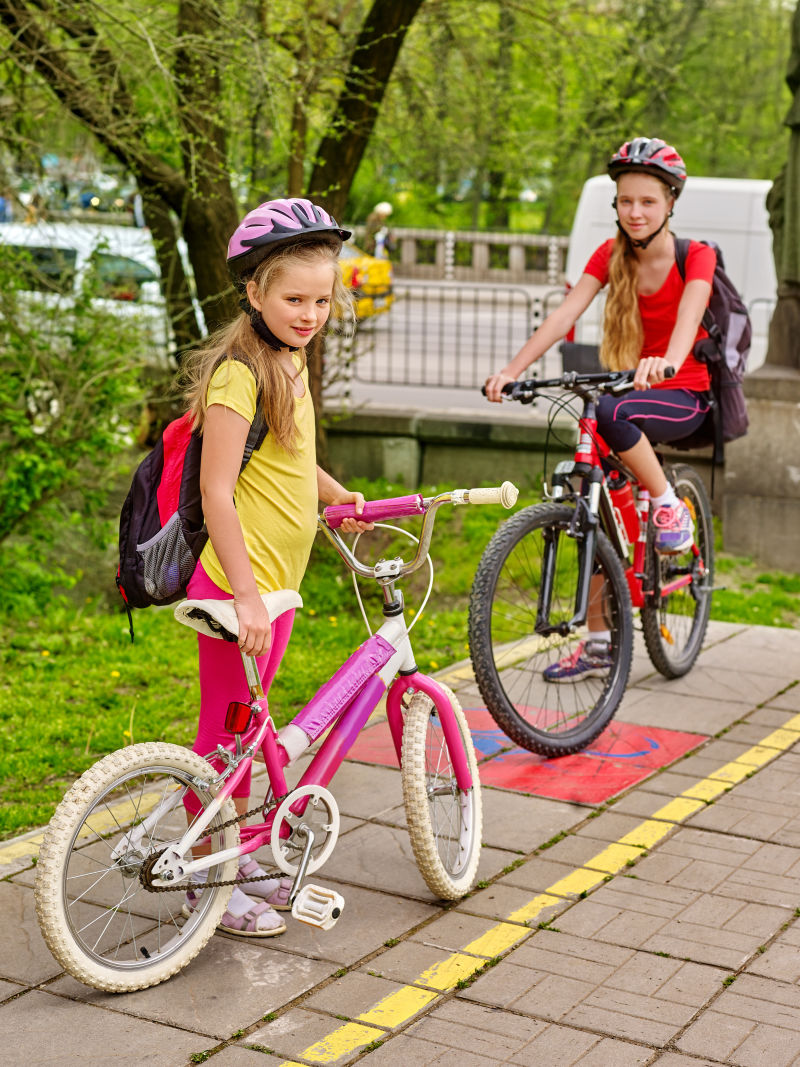 骑自行车的女孩们骑自行车的女孩女孩们戴着自行车头盔背着帆布包骑着自行车女孩们骑自行车在城市街道的黄色自行车道上与树会合