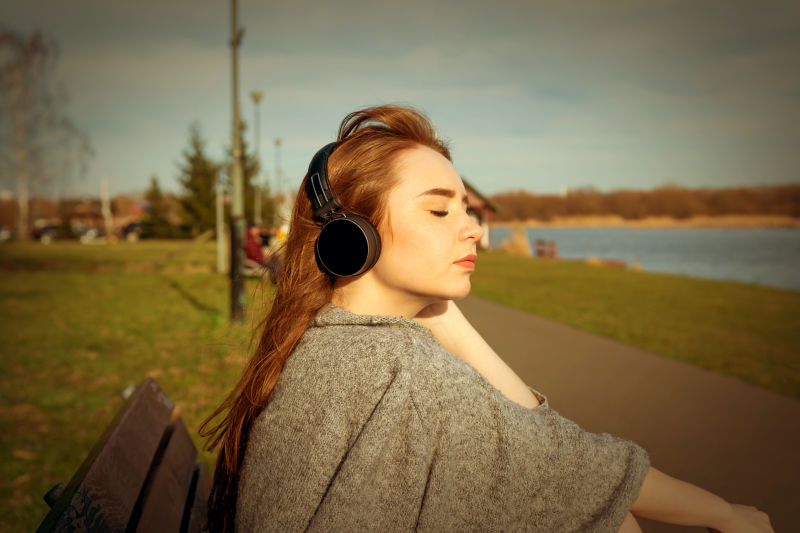 年轻快乐的红头发女孩春天在河边的公园里通过无线蓝牙耳机听音乐
