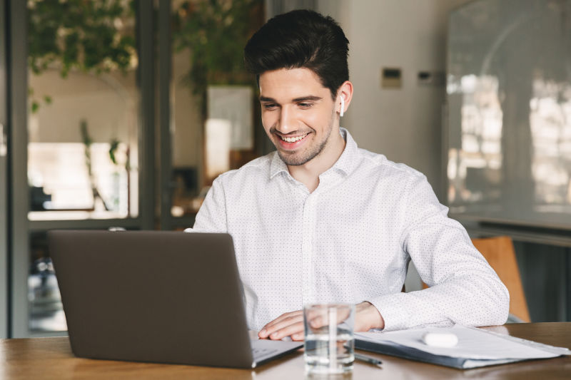一个英俊的商人穿着白衬衫戴着现代耳机坐在办公室的桌子旁看着笔记本电脑微笑着