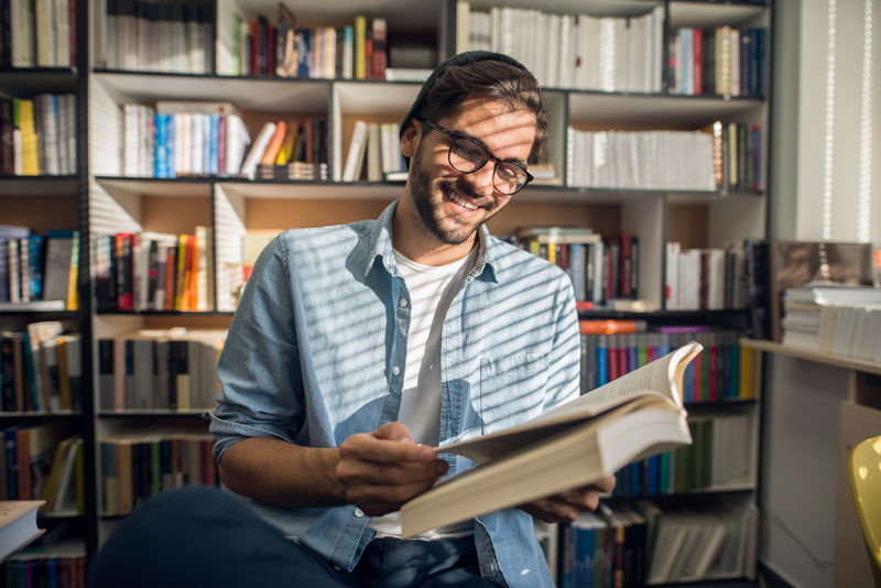 快乐的男学生坐在图书馆看书在背景书架上