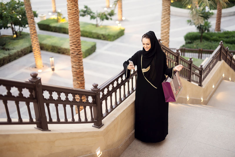 穆斯林妇女拿着购物袋和智能手机键入消息