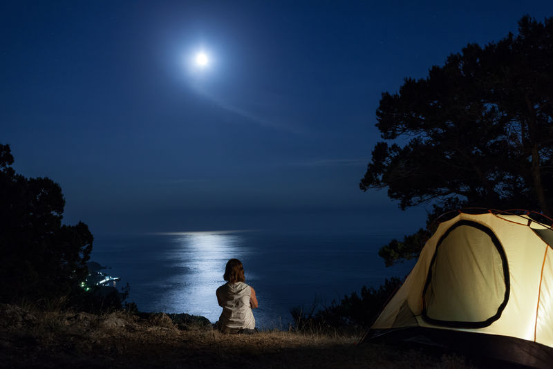 妇女在灯火通明的帐篷旁的侧影-看着海上的月亮倒影