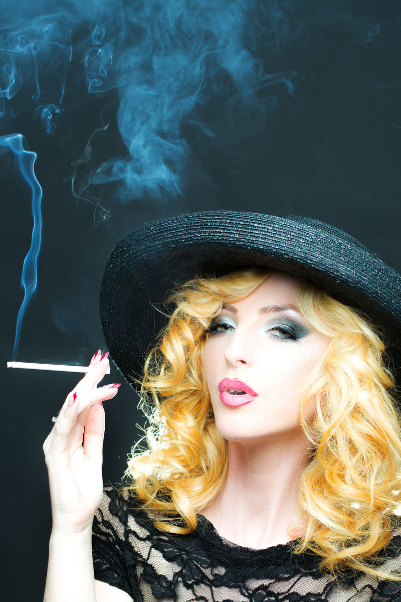 一位美丽性感的年轻复古金发女郎的肖像卷发红嘴唇戴着黑色圆帽期待在工作室背景下吸烟垂直图片