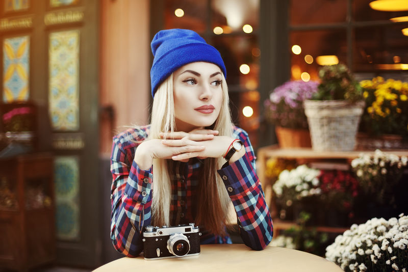 美丽时髦的年轻女子坐在街边咖啡馆喝咖啡的照片旧复古相机的时髦模特往旁边看灯光和花卉背景城市生活方式色调的