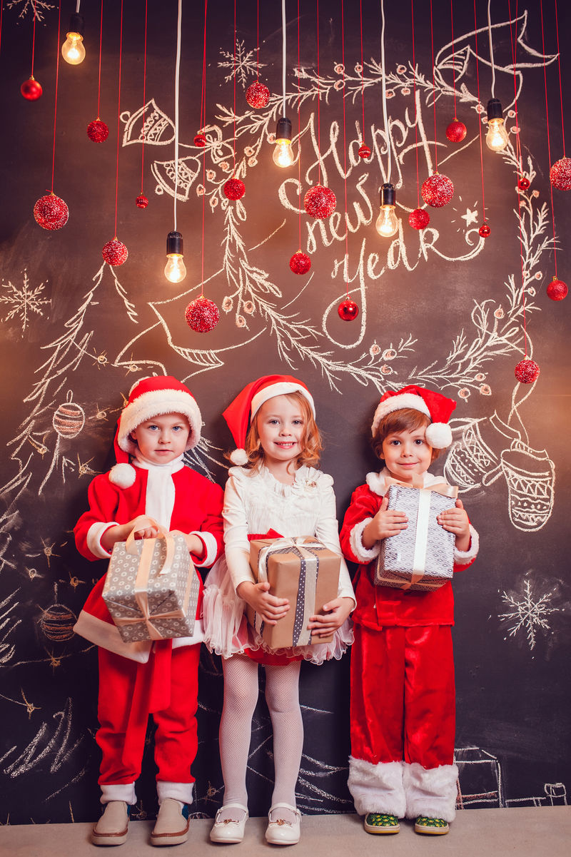 孩子们穿着圣诞老人的服装站在黑暗的背景下拿着圣诞图案的礼物新年