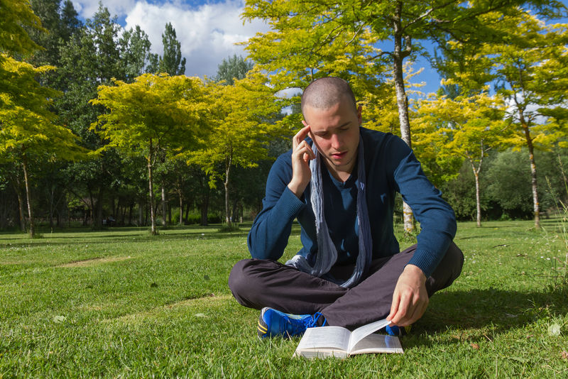一个剪发的年轻人盘腿坐在公园的草地上-读着一本体贴的书