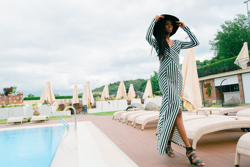 迷人美丽温柔美丽的非洲妇女头戴黑帽子穿着条纹连衣裙在欧洲豪华酒店泳池边散步的模特