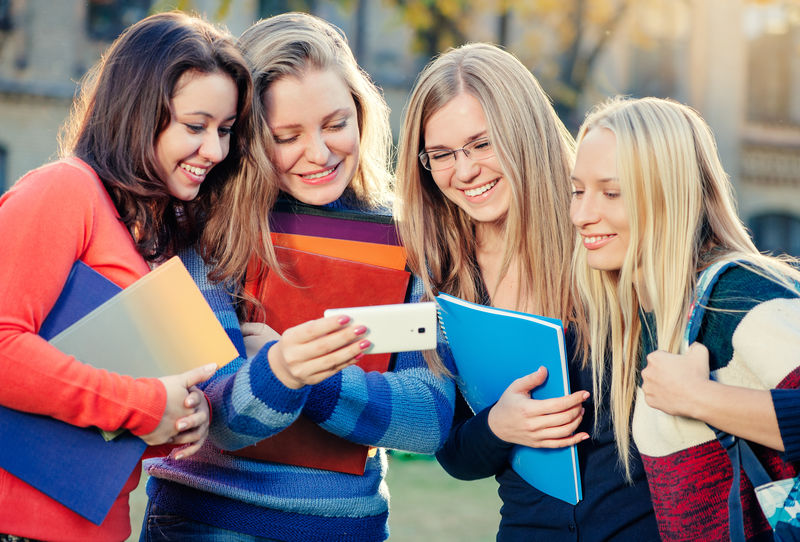 共度时光-四个微笑的女学生站在校园里讨论事情-看着智能手机