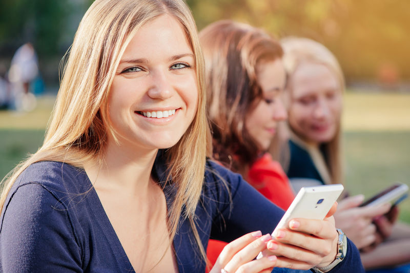与朋友分享好消息-快乐的年轻女子拿着手机-微笑着-一群人在后台交流-注意力不集中
