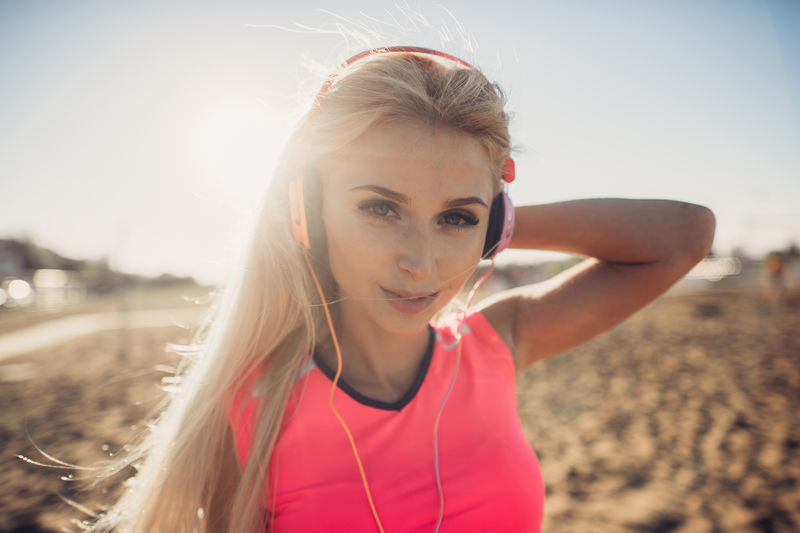 海滩上的年轻美女听音乐的肖像紧闭着笑容的金发女人脸上戴着耳机看着摄像机女孩在海滩上跑步听音乐