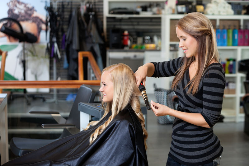 年轻造型师卷发女士的侧视图在美发沙龙呈现出新的发型