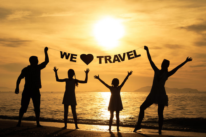 日落时分幸福的一家人站在海滩上-父母手握铭文“我们爱旅行”-友好家庭的概念