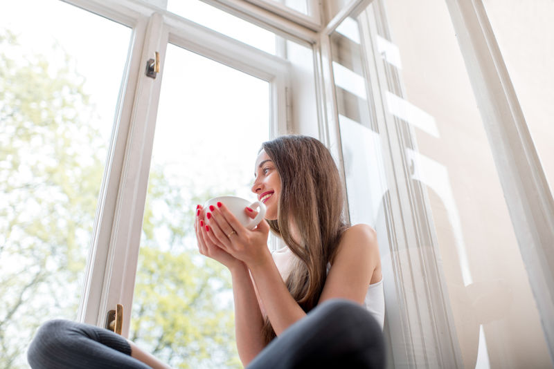 年轻可爱的女士坐在窗台上-早上拿着一杯咖啡向窗外望去