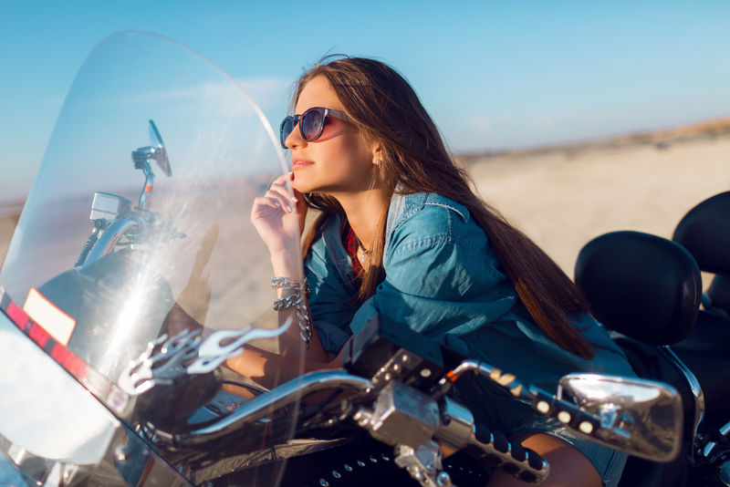 年轻迷人的性感女人坐在沙滩上的摩托车上-穿着时髦的短上衣衬衫-拥有完美的身材修长的身材和长发-户外生活写真