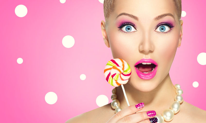 美女时尚模特吃五颜六色的棒棒糖-令人惊讶的年轻有趣的女人-粉红指甲艺术和粉红圆点背景上的化妆