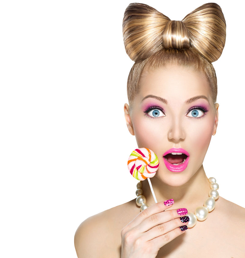 美女时尚模特吃五颜六色的棒棒糖-棒棒糖-令人惊讶的年轻风趣的女人-留着蝴蝶结的发型-粉红的指甲和白色背景的化妆品