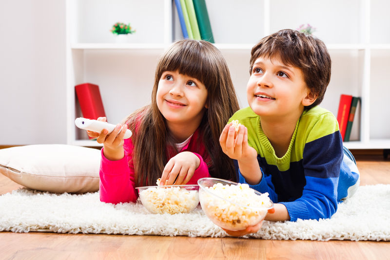 小女孩和小男孩喜欢在家吃爆米花和看电视-孩子们的休闲时间
