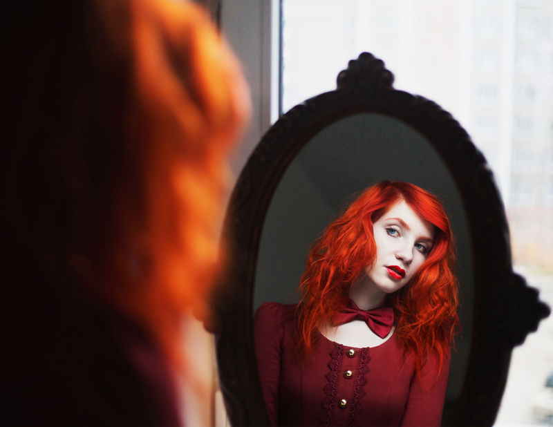 身着红色连衣裙、头上系着领结、留着红色卷发的女人在镜子里看着白色背景上的倒影。红发女孩，皮肤苍白，蓝眼睛，容光焕发，面容甜美。