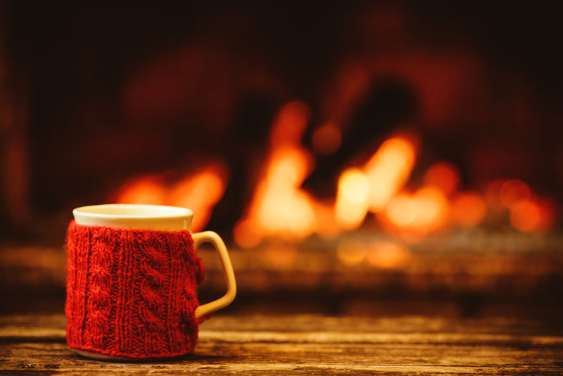 在温暖的壁炉前喝一杯热饮料-圣诞节概念-站在炉边的红色针织手套的杯子-舒适放松的神奇气氛在一个小屋