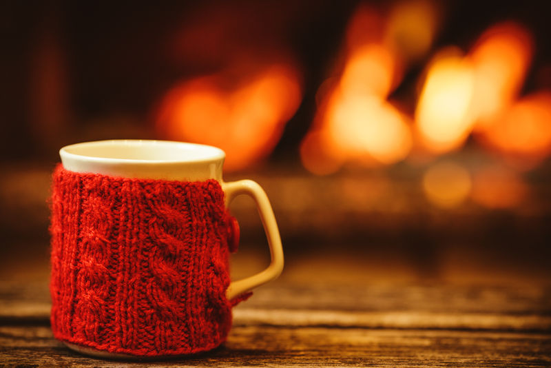 在温暖的壁炉前喝一杯热饮料-圣诞节概念-站在炉边的红色针织手套的杯子-舒适放松的神奇气氛在一个小屋