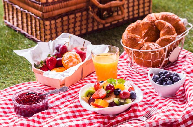 健康的素食或素食野餐-在红白相间的桌布上放上鲜果、金牛角面包、浆果果酱和热带水果沙拉-在青草上放一个篮子