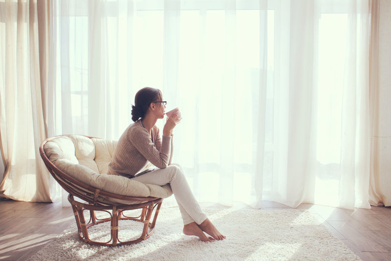 家里的年轻女子坐在窗前的现代椅子上-在客厅里休息-喝咖啡或茶
