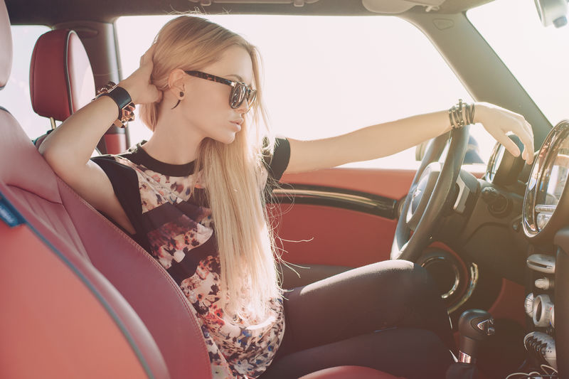 金发女郎坐在红色内饰的跑车车轮上-戴着黑色太阳镜-戴着带金属镶嵌件的皮革臂膀-手放在头旁-看着路面