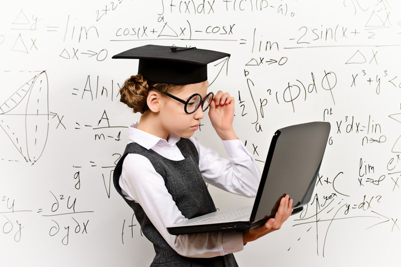戴着大眼镜、戴着学术帽的聪明女学生用笔记本电脑在黑板上完成这项任务-教育