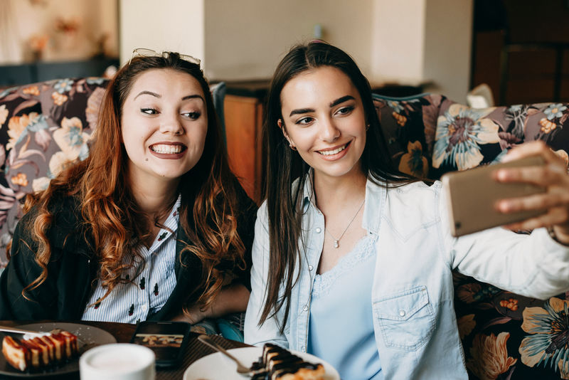 两个很棒的女朋友在咖啡店玩，一边喝咖啡一边吃蛋糕，一边用智能手机自拍。