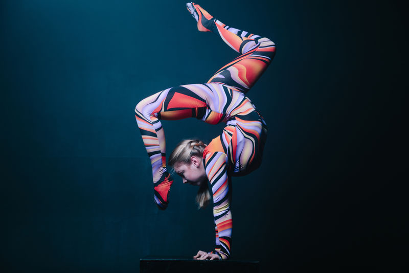 年轻柔韧的金发马戏团杂技演员在演艺厅摆姿势。在立方体上做平衡倒立。复制空间文本。