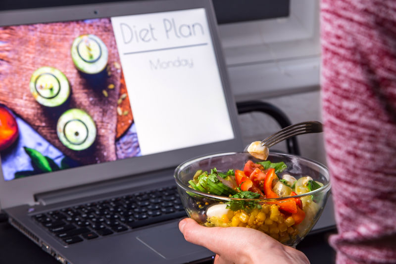 女性手中的一碗蔬菜沙拉-放在台式电脑旁-在屏幕上-计划从周一开始减肥