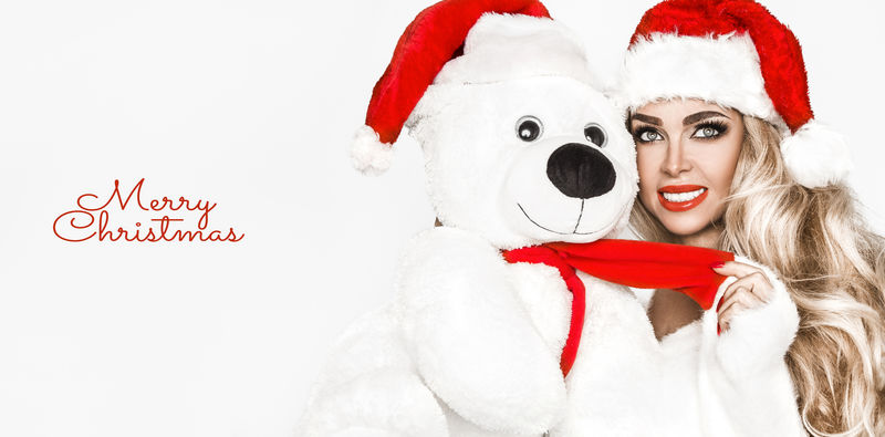 美丽的圣诞时装模特带着白色的泰迪熊。金色长直发，头戴红色圣诞帽。性感金发女青年肖像、时尚派对服装及配饰-图片