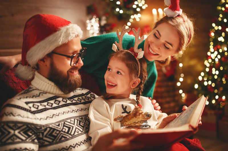 平安夜-家里的父亲、母亲和孩子在壁炉和圣诞树旁的家里读魔法书