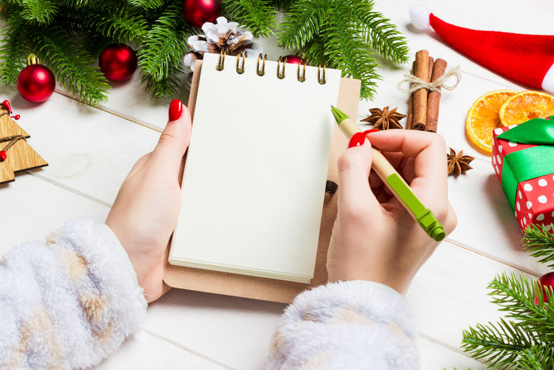 女性在木制圣诞背景笔记本上手写的俯视图。杉树和节日装饰品。愿望清单。新年概念