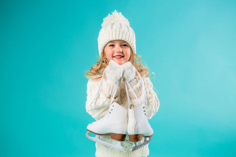 一个金发的小女孩-戴着冬天的白帽子-穿着毛衣-手里拿着溜冰鞋-微笑着-隔离在蓝色背景上-为文本留出空间-冬季运动
